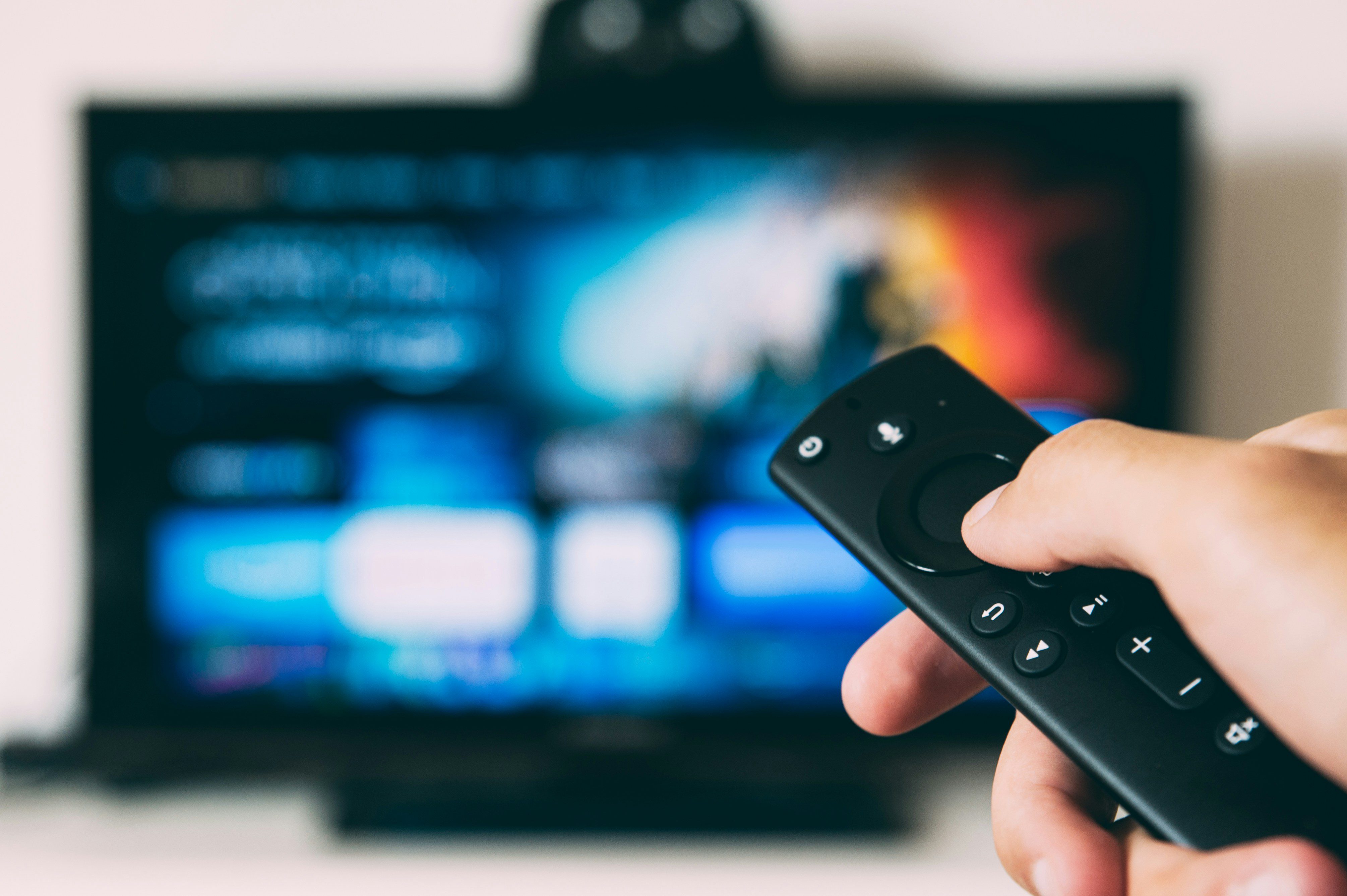 Käsi pitelee kaukosäädintä, televisio taustalla